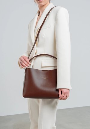 Sac à main en cuir marron avec une forme structurée, un haut plat et une fine bandoulière. Présente un logo en relief au centre et une texture lisse et brillante.