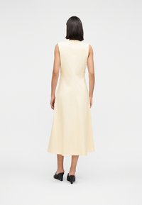 Γυναίκα που στέκεται με την πλάτη στην κάμερα, φοράει μια αμάνικη μεσομήκης κρεμ φόρεμα και μαύρα παπούτσια με τακούνι χωρίς κορδόνια, σε λευκό φόντο.