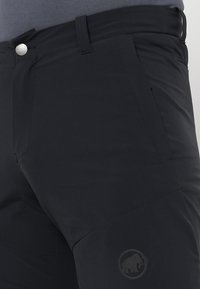 Svarta vandringsbyxor i slitstarkt tyg, med en ficka med dragkedja, bältesöglor och en logotyp nedtill på benet. Slät textur.