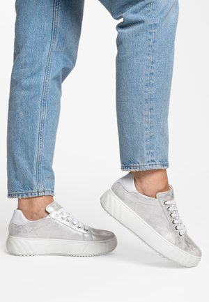 Person wearing light blue jeans and silver metallic sneakers with thick white soles, one foot raised against a white background.