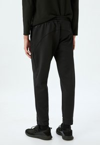 Sorte joggers med en glat tekstur, elastisk talje og tætsiddende ben. Bemærkelsesværdig syning på ryggen. Sammen med sorte sneakers.
