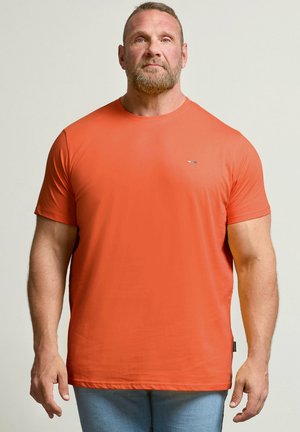 Uomo barbuto in piedi rivolto in avanti, indossa una t-shirt arancione a maniche corte e jeans azzurri su uno sfondo chiaro e semplice.