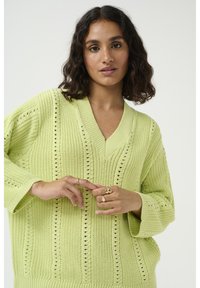 Pull en maille vert clair oversize avec un col en V, des motifs texturés horizontaux et verticaux, et des manches à épaule tombante.