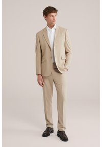 Junger Mann in beigem Anzug mit weißem Hemd und dunkelbraunen Lederschuhen, steht mit einer Hand in der Tasche vor neutralem Hintergrund.