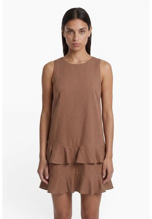 Femme portant une robe marron sans manches avec un ourlet à volants superposés, debout devant un fond blanc uni.