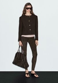 Brown cropped jacket with gold buttons, pastel pink top, dark brown suede pants, oversized brown handbag, and open-toe sandals.