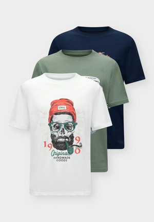 Jeune homme portant un t-shirt blanc avec un motif graphique, un short noir, des baskets blanches, tenant une veste marron devant un fond clair uni.
