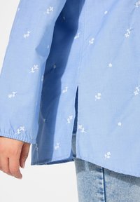 Blusa azzurra chiara con ricami floreali bianchi, maniche lunghe e spacco laterale. Il tessuto appare liscio con una consistenza leggera.