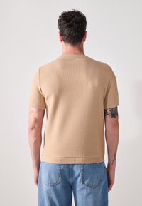 Beige short-sleeve shirt with a textured waffle pattern, round neckline, and a straight cut. Worn with light blue denim pants.