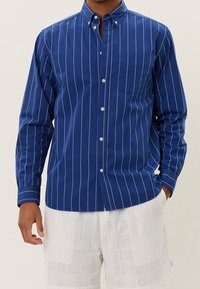 Niebieska, w paski koszula z guzikami, z kołnierzykiem, długimi rękawami i jedną kieszenią na piersi. Materiał wydaje się gładki i ma teksturę podobną do bawełny.