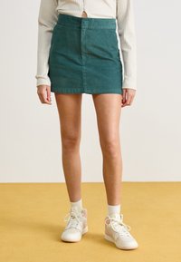 Πράσινη κοτλέ μίνι φούστα με μπροστινή ραφή, φορεμένη με ριγωτό μακρυμάνικο τοπ και λευκά αθλητικά παπούτσια με ροζ λεπτομέρειες.