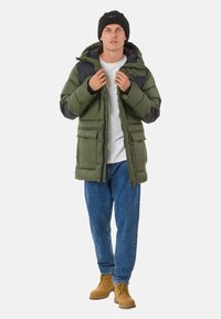 Green puffer jacket with dark gray accents, hood, and multiple front pockets. Worn over a white shirt, paired with blue jeans and tan boots.