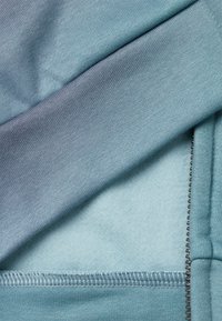 Tejido azul claro superpuesto con un material azul más oscuro. Presenta una textura suave con costuras visibles a lo largo de los bordes y un detalle de cremallera.
