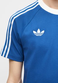Μπλε T-shirt Adidas με λευκό σχεδιασμό τριών ρίγων στους ώμους και λευκό λογότυπο τριφύλλι στο στήθος, φορεμένο από ένα άτομο.