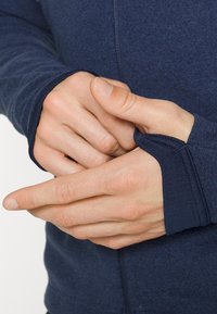 Marineblå zip-up jakke med lange ermer, med tommelhull. Mykt, teksturert stoff med sømdetaljer langs ermene og kroppen.