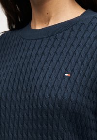 Pull en maille torsadée bleu marine porté par une personne, avec un petit logo rectangulaire rouge, blanc et bleu sur la poitrine.