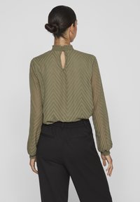 Blouse vert olive, texturée, avec un col haut, un dos en forme de clé, et des manches longues. Associée à un pantalon noir, ajusté.