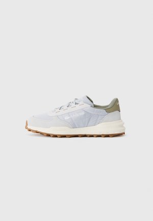 Baskets Lacoste gris clair avec semelle blanche, bande de roulement en caoutchouc naturel, et col de talon vert olive, vues de côté droit sur un fond uni.