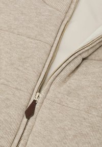 Beige gerippter Strickstoff mit teilweise geöffnetem goldfarbenem Reißverschluss an einem Kleidungsstück, der einen braunen Leder-Zippergriff besitzt.