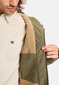 Olivegroene buitenjas met een beige, fluffy binnenvoering; met rits, voorzien van een zijzak en een corduroy shirt eronder.