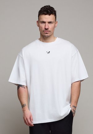 Mann mit kurzen dunklen Haaren, der ein übergroßes weißes T-Shirt mit kleinem gekreuztem Paddel-Design trägt und vor grauem Hintergrund steht.