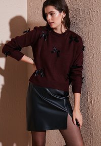 Bordeauxfarbener Strickpullover mit schwarzen Bandverzierungen und Dreiviertelärmeln, kombiniert mit einem eleganten Rock aus schwarzem Leder.