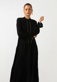 Mustapuku pitkähihainen mekko, jossa on korkea kaulus, vetoketjukiinnitys ja pörröiset hihat. Sisältää kerroksellisen hameen muotoilun ja hienovaraisen kontrastipipauksen.