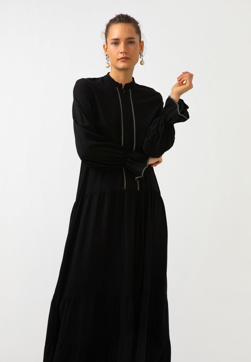 Mustapuku pitkähihainen mekko, jossa on korkea kaulus, vetoketjukiinnitys ja pörröiset hihat. Sisältää kerroksellisen hameen muotoilun ja hienovaraisen kontrastipipauksen.