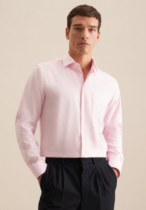Homme portant une chemise rose clair à manches longues avec une poche poitrine et un pantalon noir, debout les mains dans les poches.
