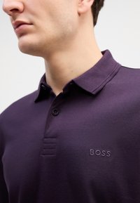Ljubičasta polo majica s ovratnikom, s dva gumba i stiklanim "BOSS" logom na lijevoj strani prsnog koša. Glatka textura i čist dizajn.