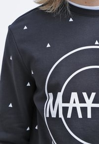 Felpa nera con motivi triangolari bianchi e testo grafico "MAY." Presenta un design con logo rotondo. Realizzata in un tessuto soffice e strutturato.