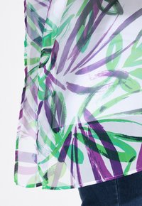 Gennemsigtig bluse med et farverigt botanisk mønster i grøn og lilla på en hvid baggrund, lavet af let stof og med et svævende design.