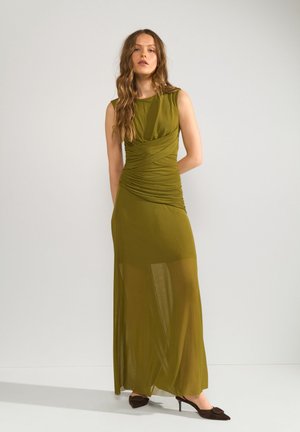 Femme aux longs cheveux ondulés portant une robe longue sans manches vert olive avec des détails froncés et des talons pointus noirs.