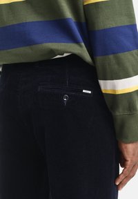 Maglione verde con strisce orizzontali gialle, blu e bianche. Pantaloni in corduroy blu scuro con tasca posteriore e dettaglio con bottone.