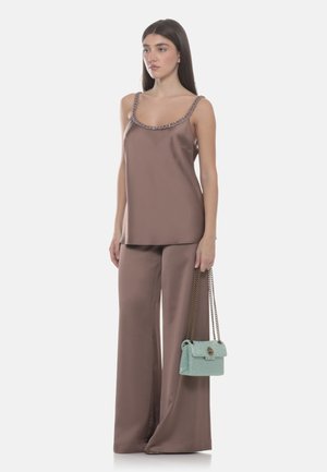 Femme portant un haut sans manches en satin mauve et un pantalon à jambes larges, tenant un sac à bandoulière en chaîne bleu clair, debout devant un fond uni.