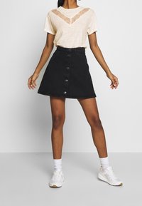 Mujer con una blusa corta de mangas, beige y con encaje, una falda negra con botones al frente, calcetines blancos y zapatillas blancas, de pie contra un fondo blanco.