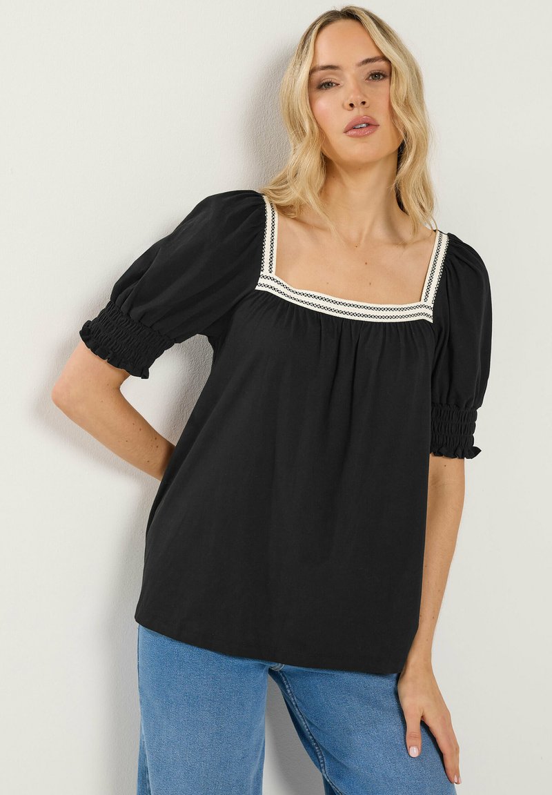 Mujer con cabello rubio que lleva una blusa negra de cuello cuadrado con mangas abullonadas y borde de encaje blanco, combinada con jeans azules, de pie contra una pared blanca.