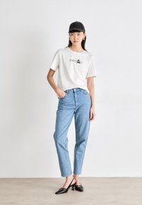 Bijela majica s logom, svijetloplave traperice i crne cipele s visokom petom. Outfit je upotpunjen crnom kapom i minimalnim dodacima.