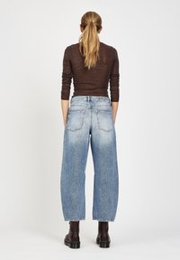 Frau mit langem blonden Pferdeschwanz, die einen braunen Langarmtop, hellblaue weite Jeans und dunkelbraune Stiefeletten trägt, steht mit dem Rücken zur Kamera.