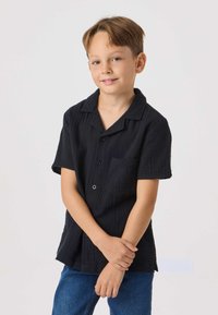 Sort kortærmet skjorte med tekstureret stof, knaplukning foran og brystlomme, kombineret med blå denim-jeans.