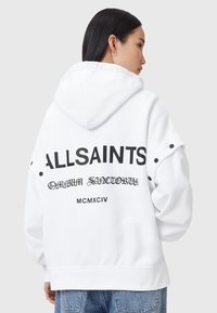 Sudadera blanca con texto negro que dice "ALLSAINTS" y "MCMXCIV" en la parte posterior. Incluye detalles de hardware y un corte relajado.