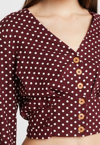 Blouse bordeaux à pois avec un décolleté en V profond, un détail froncé et des boutons en bois sur le devant. Confectionnée en tissu léger.