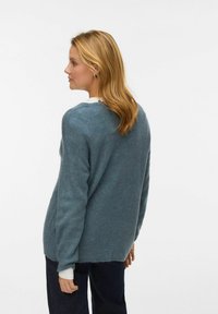 Adīts zilas krāsas džemperis ar ribotu tekstūru, apaļu kakla izgriezumu un nolaistām pleciem, valkāts kopā ar tumšiem džinsiem, redzams no aizmugures.