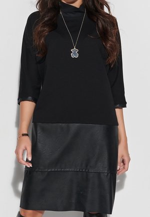 Woman wearing a black dress with three-quarter sleeves and leather-look skirt, accessorized with a bear-shaped pendant necklace.