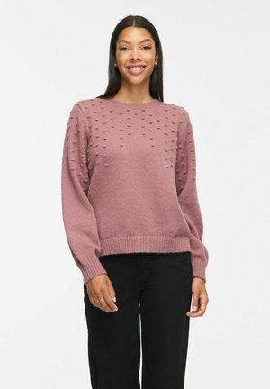 Femme aux longs cheveux foncés portant un pull texturé mauve et un pantalon noir, souriante et debout sur un fond blanc uni.