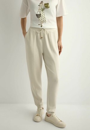 Osoba ubrana w białą koszulkę z grafiką kobiety, beżowe spodnie dresowe ze ściągaczem oraz dopasowane beżowe trampki, stojąca z rękami w kieszeniach.
