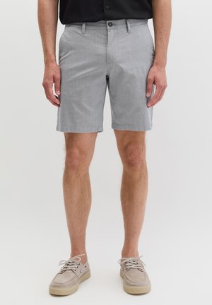 Man draagt lichtgrijze op maat gemaakte korte broek, beige veterschoenen en een zwart overhemd, staand tegen een effen achtergrond.