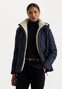 Námornícky prešívaný kabát s límcom z umelého kožušiny, so zipsom vpredu a kontrastnými hnedými akcentmi. Skombinovaný s čiernym rolákom a opaskom.