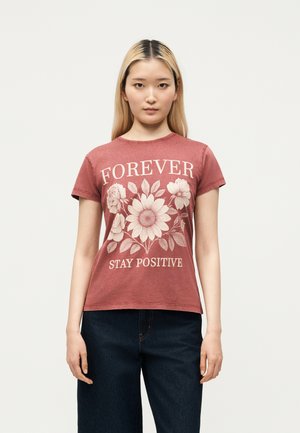 Femme aux longs cheveux blonds portant un t-shirt rouge délavé avec un motif floral et le texte « FOREVER STAY POSITIVE », associé à un jean foncé.