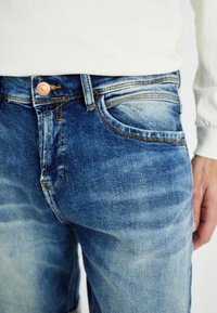 Jean en denim bleu moyen avec un délavage, comportant un bouton en métal cuivré, des coutures orange et des poches avant. Texture lisse.